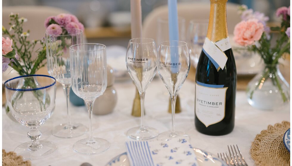 A beautiful pastel blue and pink tablescape design with pattern table linen, brass homeware accessories and  a bottle of Nyetimber