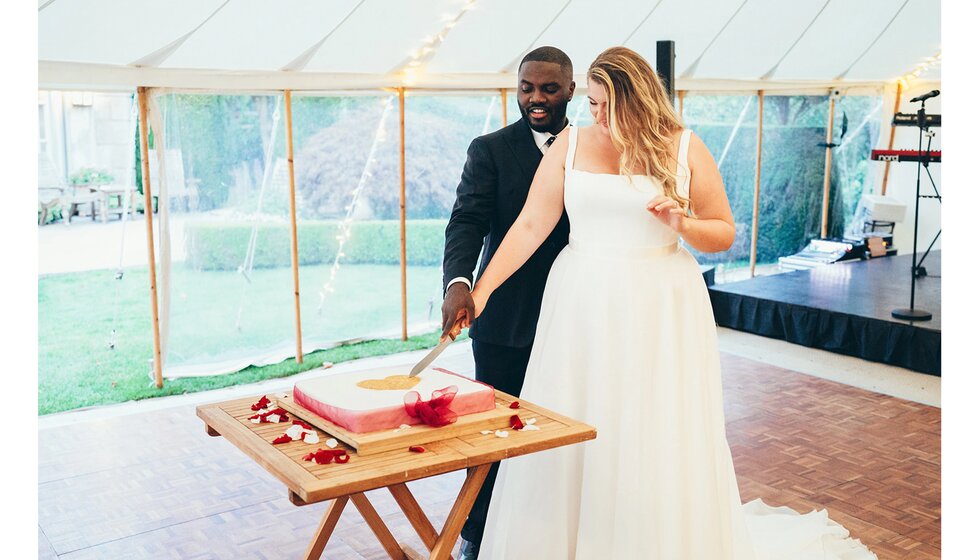 The bride & groom cut a wedding cake decorated with a heart and red ribbon.