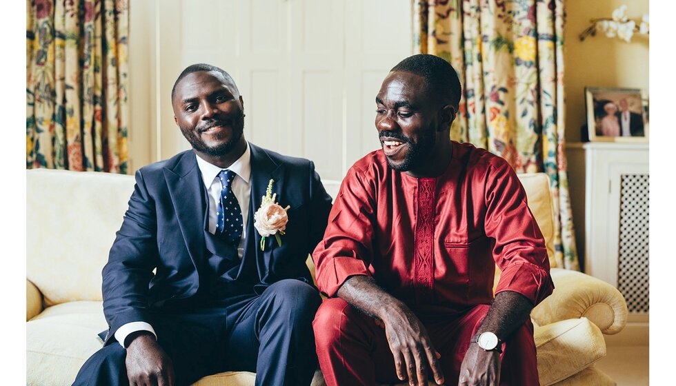 The groom in a dark navy suit sits with his groomsman who wears a traditional red Nigerian celebration suit