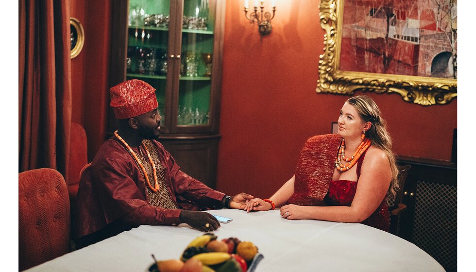 The bride and groom sit behind the table, wearing traditional red Nigerian wedding outfits