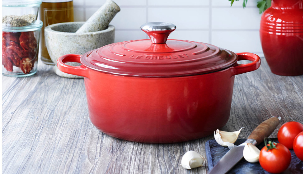 An image of a cerise round cast iron with tomatoes and garlic in the foreground on a chopping board.