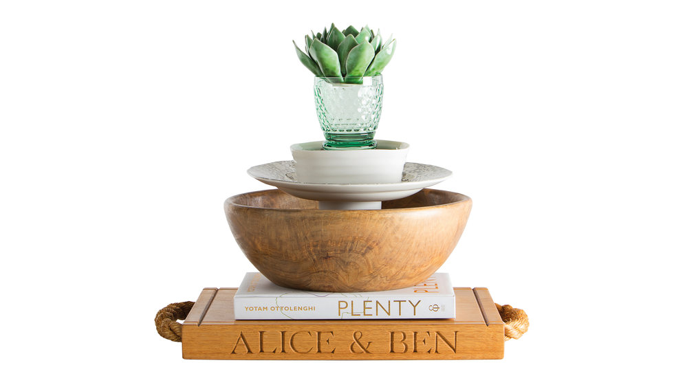  A stack of wedding presents including an engraved wooden chopping board, a cook book, a mango wood salad bowl, a butter dish a glass and a tea light.