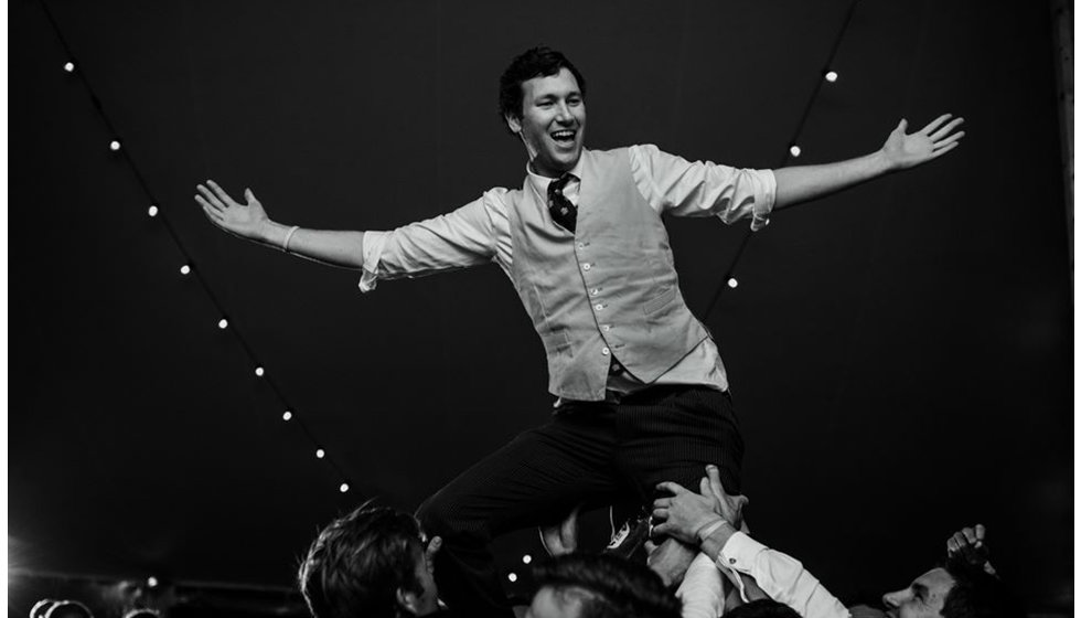 Groom Sam on the shoulders of a guest at his wedding on the dance-floor in his marquee wearing a smart waistcoat. 