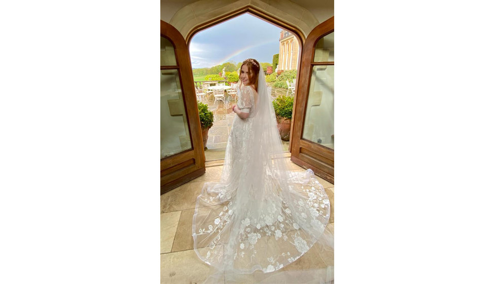 Sarah holds her bouquet wearing her wedding dress, in front of an open door with a rainbow in the background.