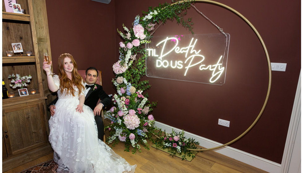 Sarah and Nico pose under a neon sign that says 'Til Death do us Party'. 