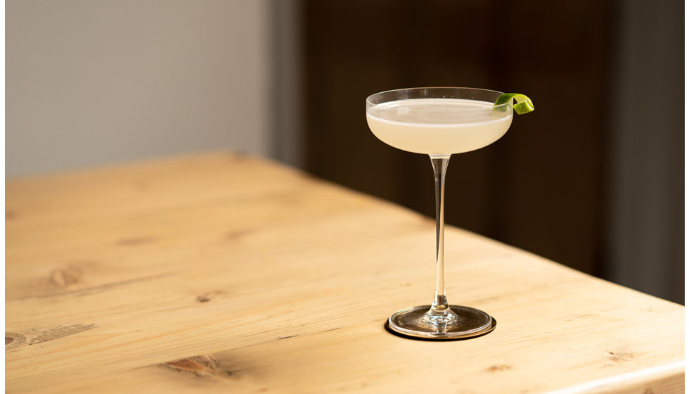 A classic cocktail by The Cocktail Society in a coupe glass on a table.