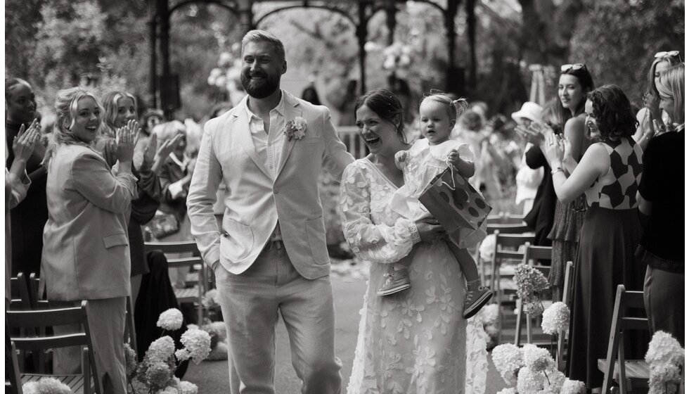 Whimsical Floral Wedding in London Park | Wedding guests are standing and clapping for a just-married couple who is walking down the aisle after their wedding ceremony, holding their small daughter, and smiling.