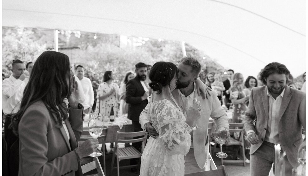 Whimsical Floral Wedding in London Park| The bride and groom are holding a glass of wine and kissing after the wedding toast.