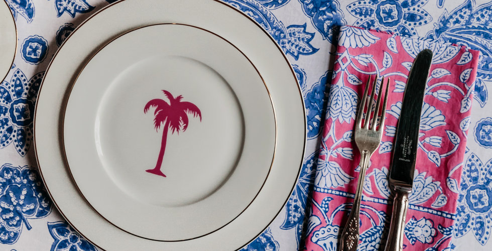 An Alice Peto pink palm tree side plate on top of a gold rimmed dinner plate on top of a blue and white floral tablecloth with a pink napkin and silver cutlery.